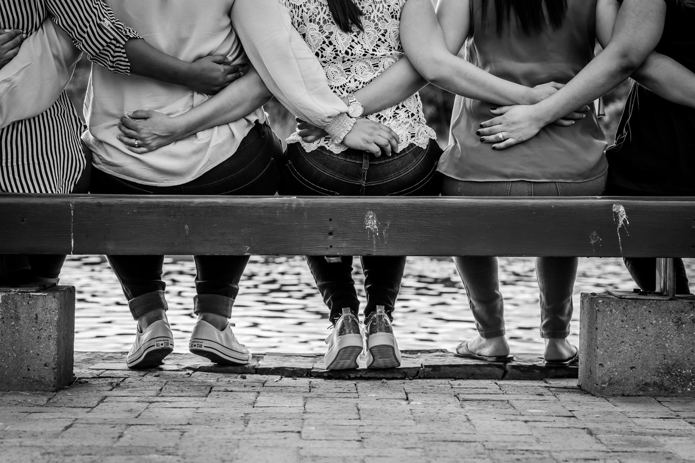 Friends sitting side by side on a bench by the water, arms around each other.