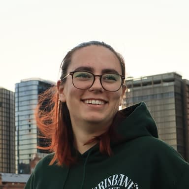 Reynae smiling in front of city buildings, wearing glasses and a green Brisbane Bike Girlies hoodie.